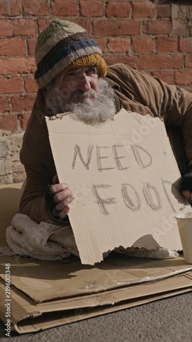 Vertical video of senior Caucasian homeless man sitting on sidewalk holding cardboard sign begging for help. Clip serves poverty awareness, charity campaigns and social welfare initiatives