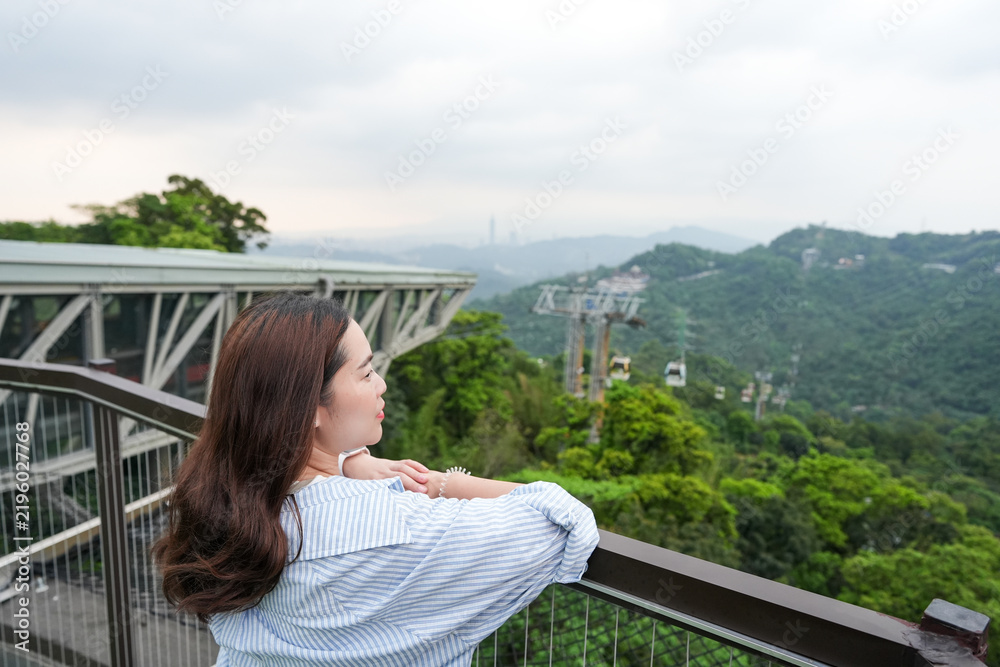 Obraz premium A side-profile shot of a young Asian woman leaning against a railing at an elevated observation platform, taking in the panoramic landscape of green hills and the Maokong Gondola cable car line in Tai