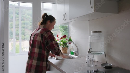 Woman washes a wooden cutting board with a sponge and running water at the kitchen sink in a modern apartment.