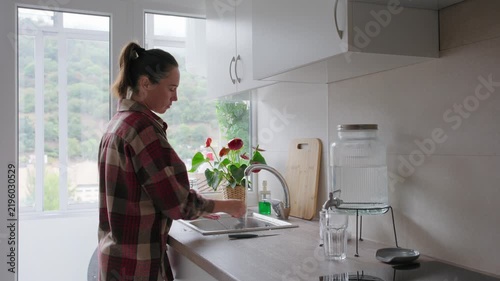 Woman washes kitchen knives under running tap water and dries them with a dish towel in modern bright kitchen.