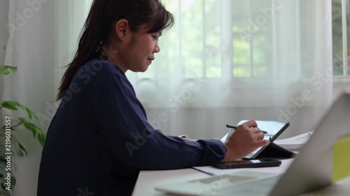 Focused woman drafting strategy document on slim tablet device with pen stylus while seated near window inside peaceful corporate office room during daytime working hours