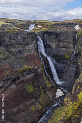 Granni waterfall on the Fossa River in Thjorsardalur, Iceland, bends down layered cliffs into a deep gorge, with snow patches, green vegetation and a winding stream below the stepped falls.