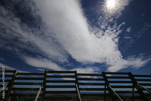 Vue de face d'une longue barrière en bois avec des traverses horizontales implantée en zone rurale montagneuse. Longs nuages blancs emportés par le vent dans un ciel bleu d'été.