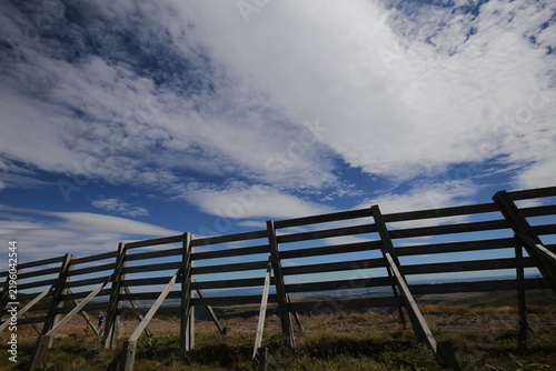 Vue de face d'une longue barrière en bois avec des traverses horizontales implantée en zone rurale montagneuse. Longs nuages blancs emportés par le vent dans un ciel bleu d'été.