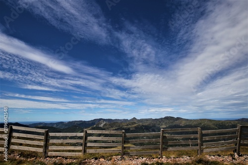 Vue de face d'une longue barrière en bois avec des traverses horizontales implantée en zone rurale montagneuse. Longs nuages blancs emportés par le vent dans un ciel bleu d'été.