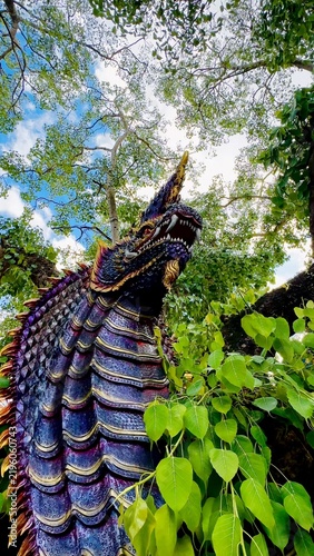 Colorful Naga serpent sculpture beside a large tree in a lush outdoor setting, with intricate scales and vivid details surrounded by green foliage and bright blue sky.