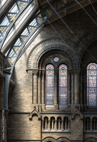 Sunlight streams through the arched windows of a building, creating distinct patterns of light and shadow on the detailed stonework walls, arched windows and stained-glass in the Natural History Museu