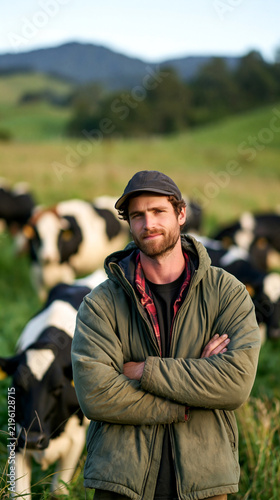Confident farmer stands arms crossed among grazing dairy cows in a lush green pasture, morning light and distant hills capturing authentic rural life.
