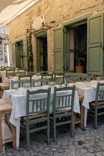 Rustic Outdoor Café with Sage Green Chairs and Gingham Tablecloths in Mediterranean Coastal Town. Alacati, Turkiye. Traditional taverna in Aegean with vintage green shutters, white tablecloths. 