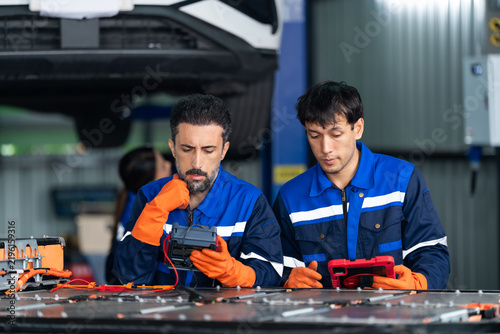 Two adult male EV technicians review battery diagnostics using multimeter and rugged tablet beside exposed high voltage battery pack in modern automotive workshop, supporting teamwork and inspection.