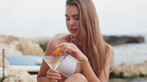 Model enjoys a cocktail while relaxing by the shore near the ocean