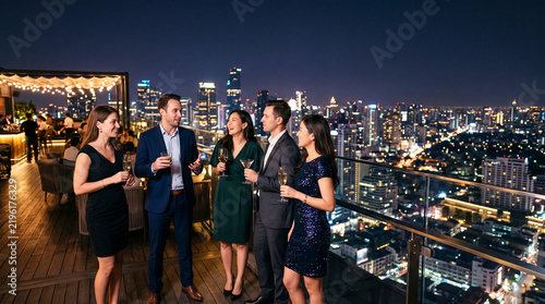 Group of colleagues celebrating and enjoying drinks at a rooftop bar overlooking the illuminated city skyline at night.