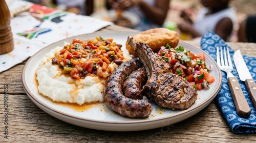 Traditional South African braai plate with boerewors, lamb chop, pap, and chakalaka. Heritage Day food celebration on wooden table
