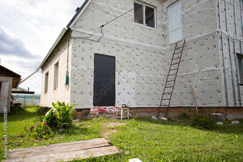 unfinished residential house facade covered with exterior insulation membrane and fasteners, with ladder and entrance door showing ongoing energy retrofit and renovation