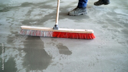 Worker in safety boots sweeping wet concrete floor with large red push broom at construction site