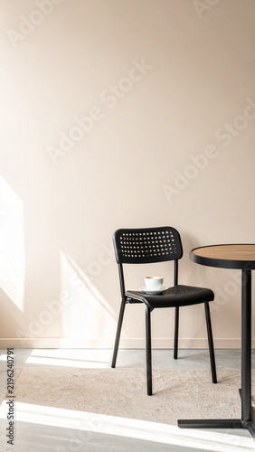 A modern black wooden chair and a wooden table with a cup on it placed on a rug in a minimalist room with sunlight streaming across the wall.