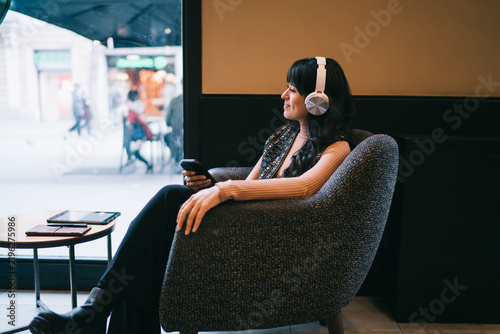 Caucasian female student wearing wireless headphones while sitting in cafe armchair with smartphone, browsing e-commerce retail store discount sales promotional deals and audio offers
