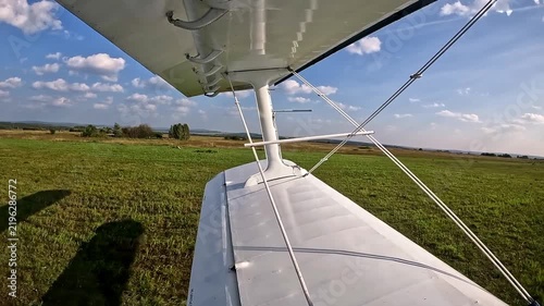 A close‑up of the wing and struts of a light aircraft on a grass airfield on a clear summer day with a cloudy blue sky.
