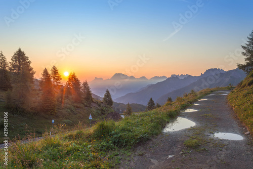 Sunrise casting golden light over dolomite peaks, reflecting in puddles on a winding mountain path