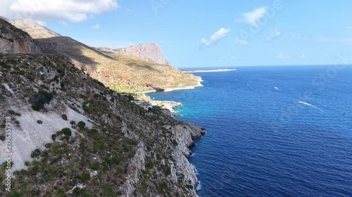 Coastal landscape view of rocky cliffs and turquoise waters, showcasing the gradual transition of shoreline and distant mountains under a bright blue sky with scattered clouds