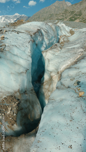 Foto Deep glacial crevasse with blue ice and rocky debris, snowcapped mountains under