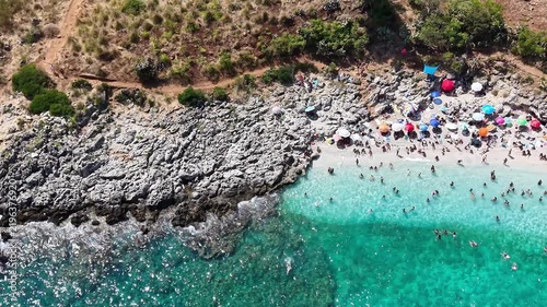 Aerial view of a vibrant beach scene with people swimming, sunbathing, and relaxing on the sandy shore, surrounded by colorful umbrellas and rocky coastline under bright sunlight. Sea resort at warm s
