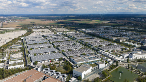 Aerial view of Messe Munchen exhibition center with large interconnected halls, flat roofs, surrounding green spaces, and a nearby pond in Munchen, Bayern, Germany.