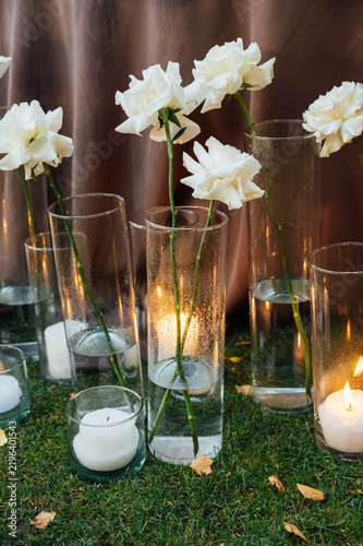 Romantic wedding floor decor with white roses in tall glass vases and burning candles on the grass.