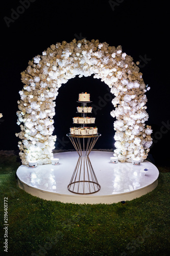 Tiered wedding cake and cupcake tower stand in front of illuminated floral arch at night.