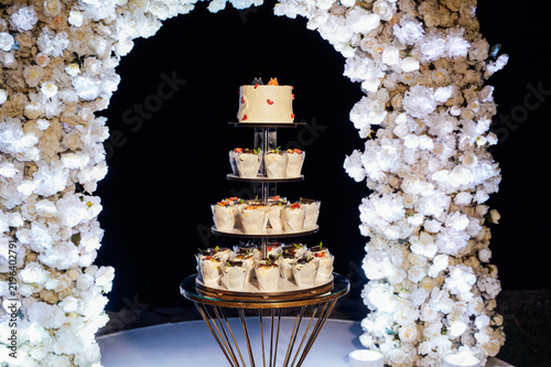 Tiered wedding cake and cupcake tower stand in front of illuminated floral arch at night.