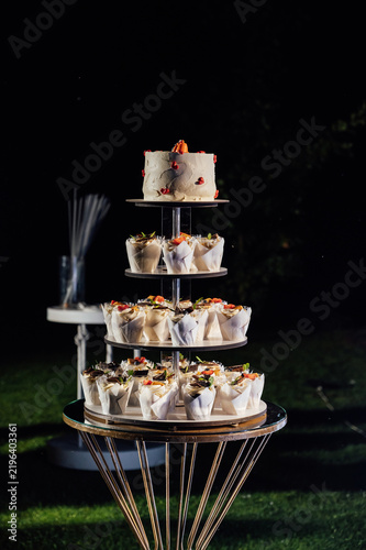Tiered wedding cake and cupcake tower stand in front of illuminated floral arch at night.