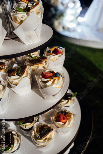 Tiered wedding cake and cupcake tower stand in front of illuminated floral arch at night.