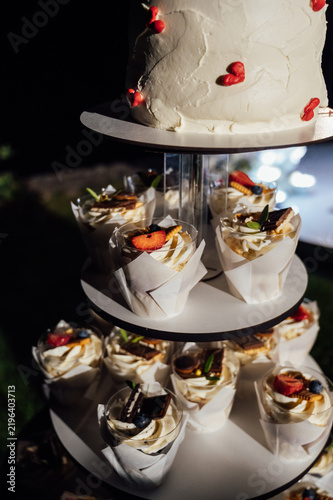 Tiered wedding cake and cupcake tower stand in front of illuminated floral arch at night.