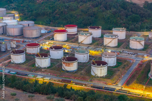 Aerial view of a fuel depot with numerous large cylindrical storage tanks, some with red tops, situated amidst green hills and industrial infrastructure in Rome, Lazio, Italy.