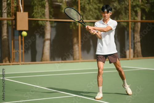 фотографія Young Asian Man Playing Tennis Outdoors, Preparing Backhand Stroke on Green Cour