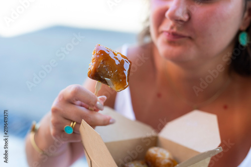 A woman holds a honey-soaked loukoumades dessert on a wooden stick in Kerkira, Greece. She eats traditional Greek fried dough pastry from a takeout box.