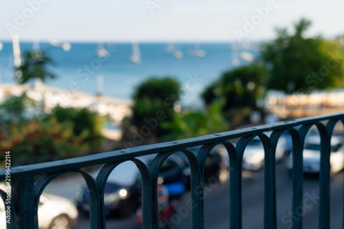 A dark metal balcony railing stands in sharp focus in Kerkira, Greece. A blurred coastal street and blue sea filled with boats extend into the background.
