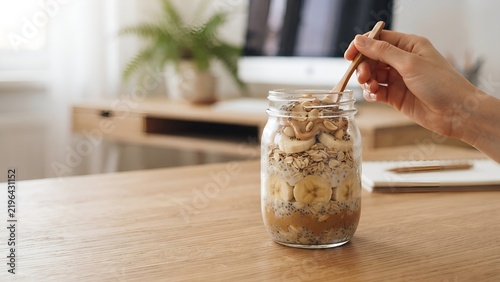 Person stirring overnight oats in a glass jar on a wooden table.