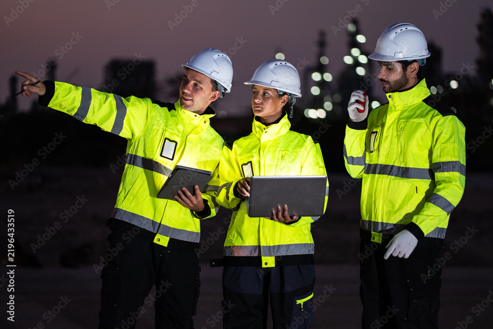 Fototapeta premium A group of industrial workers in safety gear collaborate at refinery factory at night, planning operations and reviewing data on digital devices while discussing project progress together.