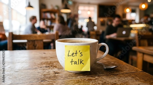 A cup of hot coffee with a yellow sticky note that says Let's Talk on a wooden table in a busy coffee shop cafe