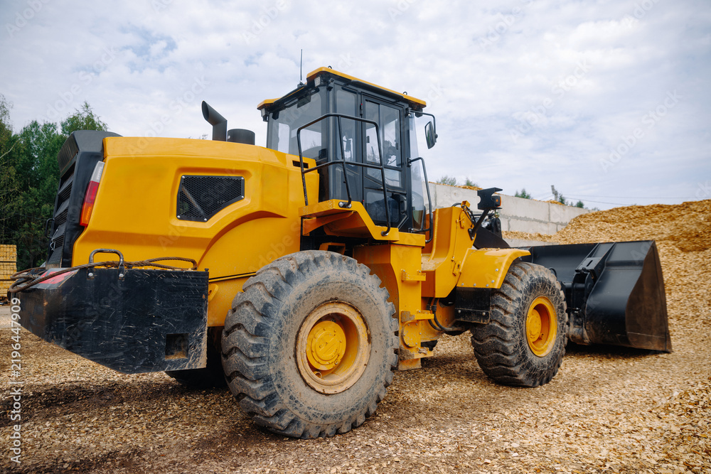Fototapeta premium Yellow wheel loader parked on gravel surface with large pile of wood chips in the background, showcasing heavy machinery in an outdoor construction site