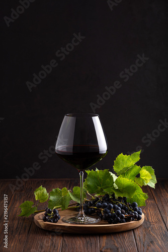 Obraz na plátně Elegant glass of rich red wine with fresh dark grapes and green leaves on rustic