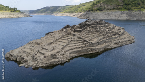 Aerial view of Castro Candaz, an ancient hillfort ruin emerging from the waters of the Mino River, surrounded by terraced hillsides in Belesar, Lugo, Spain.