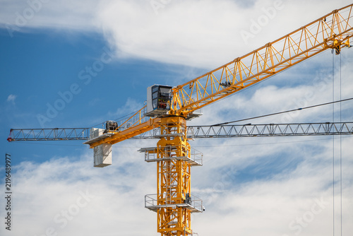 Bild auf Leinwand A bright yellow tower crane with a glazed operator cabin and counterweights extends toward a vivid blue sky with soft white clouds
