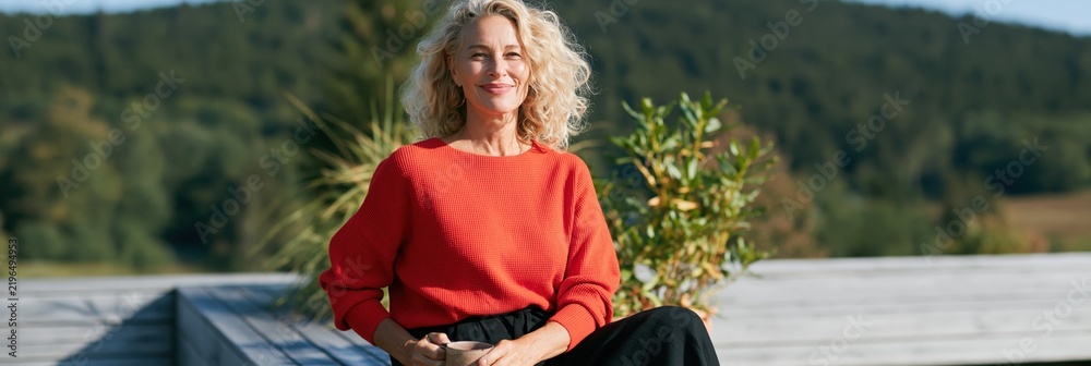 Obraz premium Smiling middle aged woman in orange sweater sitting on a wooden terrace with potted plants and mountain view for wellness portrait and lifestyle background design
