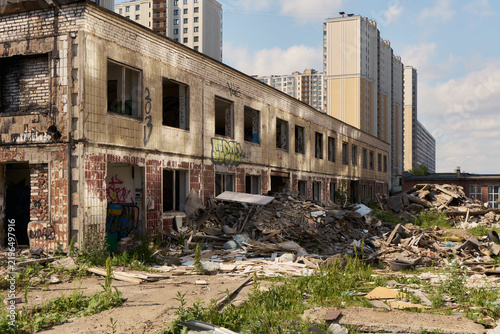Abandoned brick building with graffiti and rubble foreground, decaying walls, exposed windows, overgrowth and urban decay beside modern high-rise apartments under clear blue sky