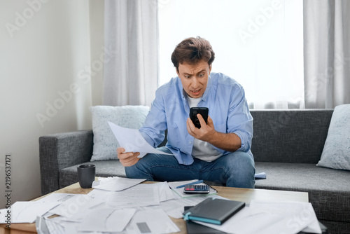 Foto Shocked man holding utility bills and smartphone, experiencing financial stress from rising living costs, inflation, and unexpected household expenses during economic crisis
