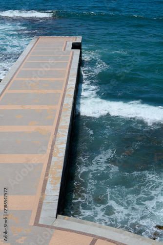 A geometric walkway structure bordered by vibrant turquoise sea water and white sea foam along the coastal edge.