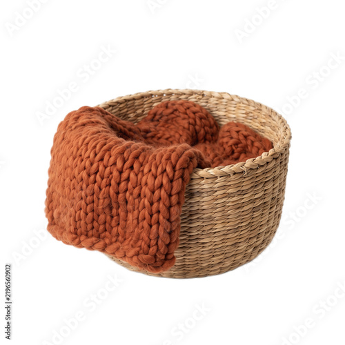 A woven straw basket containing a thick orange knitted wool blanket against a white background with chunky knit blanket color against plain with basket holds soft