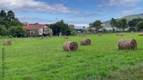 Rural landscape in Sukarrieta, Basque Country, with hay bales scattered across a green meadow, a traditional farmhouse, trees and wooded hills under a partly cloudy sky